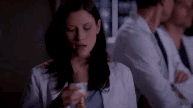 a woman in a lab coat is drinking from a cup while a group of doctors are standing behind her .