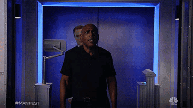 a man in a black shirt is standing in front of a blue door with nbc written on the bottom