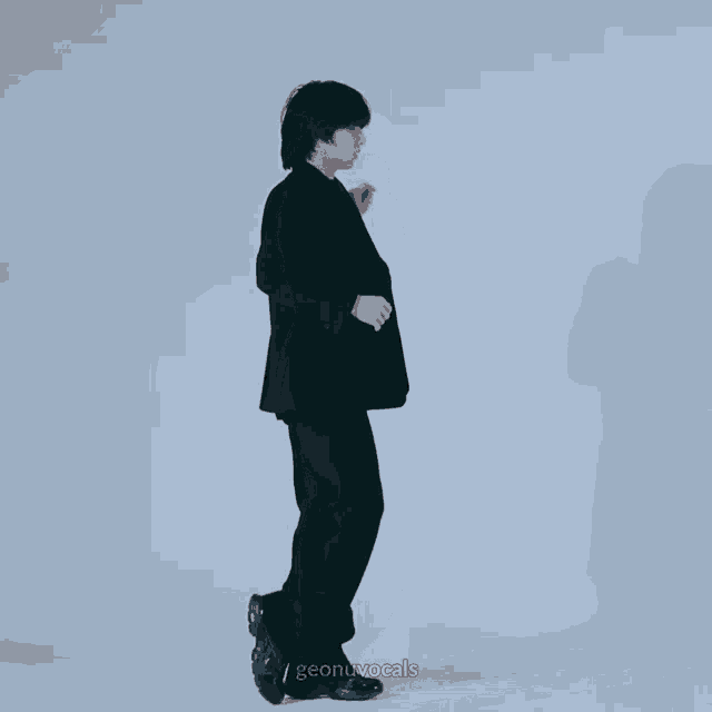 a man in a black suit is standing in front of a white wall with the words geonuvocals written on the bottom right