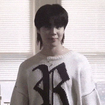 a young man wearing a white sweater with a black letter r on it is standing in front of a window .