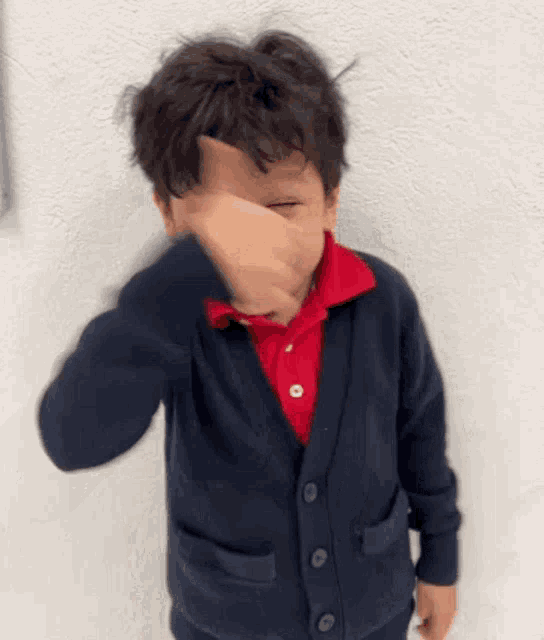 a young boy wearing a blue cardigan and a red polo shirt is covering his face with his hand .