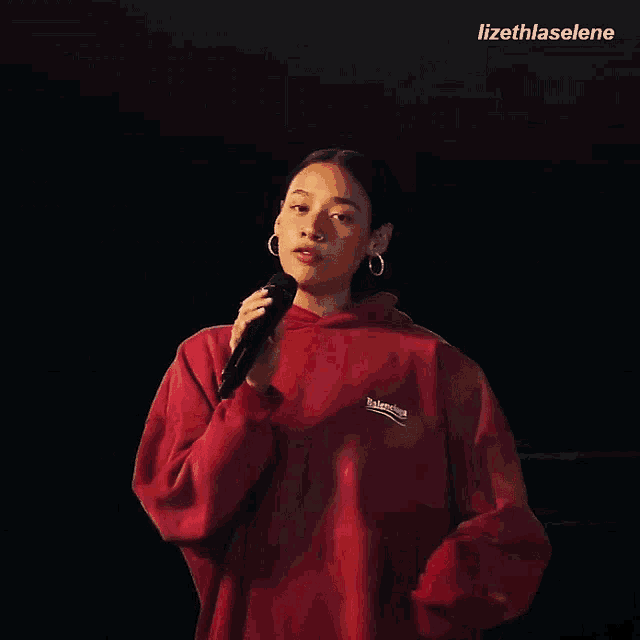 a woman singing into a microphone wearing a red balenciaga sweatshirt