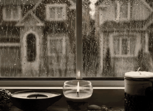 a candle is lit in front of a window with rain drops on it and a house in the background .