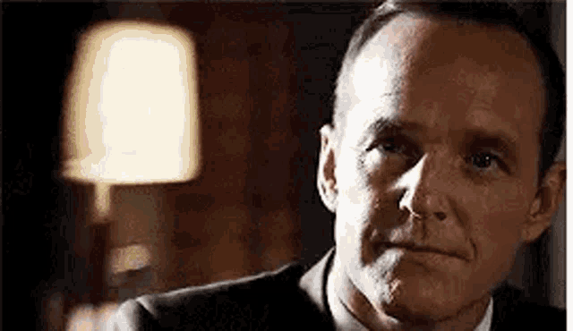 a close up of a man in a suit and tie looking at the camera with a lamp in the background .
