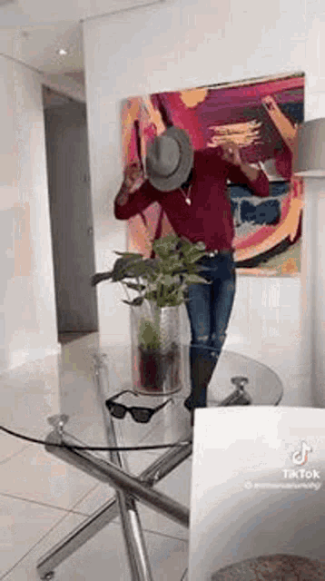 a man in a hat is standing in front of a glass table with a plant in it .