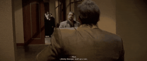 a man in a suit talks to another man in a hallway and says " hya jimmy george wait up a sec "