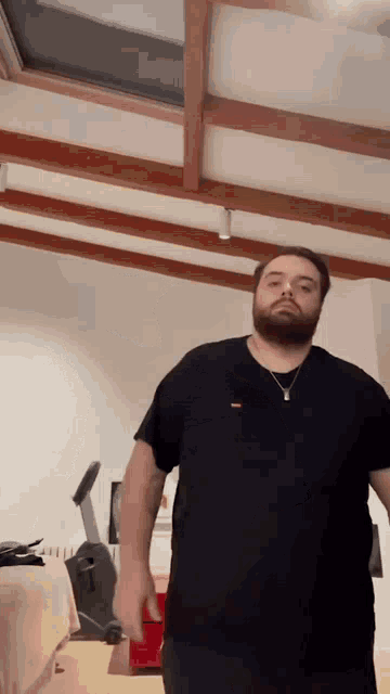 a man with a beard is standing in a room wearing a black t-shirt and a necklace .