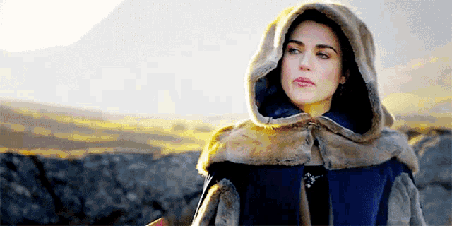 a woman wearing a fur cape with a hood is standing on a rocky hillside .