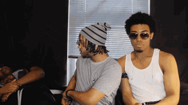 a man wearing sunglasses and a beanie sits next to another man wearing a white tank top