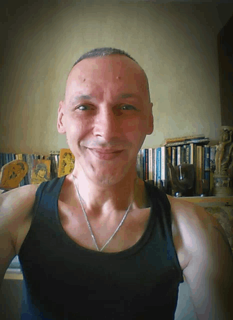 a man wearing a black tank top smiles in front of a bookshelf