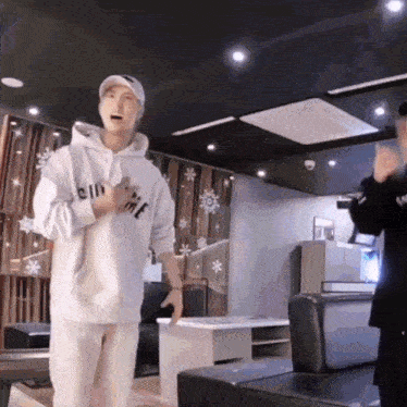 a man in a white hoodie is standing in a room with a couch .