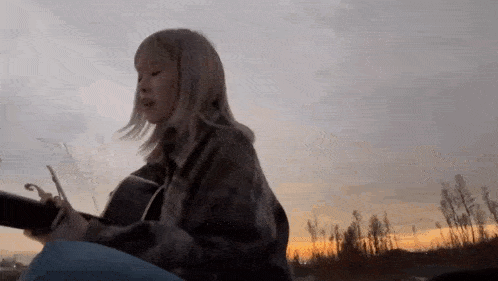 a young woman is playing an acoustic guitar in front of a sunset in the countryside .