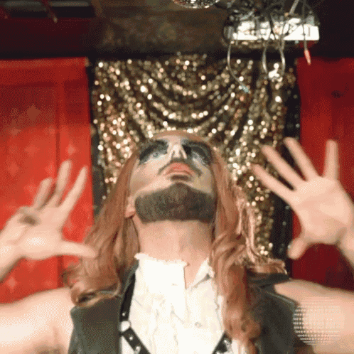 a man with a beard and makeup on his face has his hands up