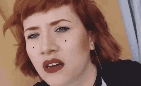 a woman with red hair and blue eyes is wearing red lipstick and earrings and making a funny face .