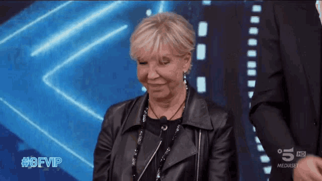 a woman in a black leather jacket stands in front of a screen that says 5 hd