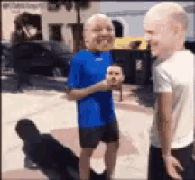 two men are standing on a sidewalk talking to each other . one of the men is wearing a blue shirt and shorts .