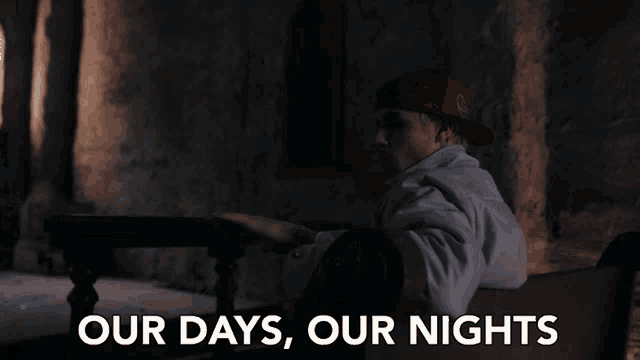 a man in a red hat sits in a dark room with the words our days our nights above him