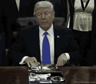 a man in a suit and tie is sitting at a table with a box on it that says donald draws.com