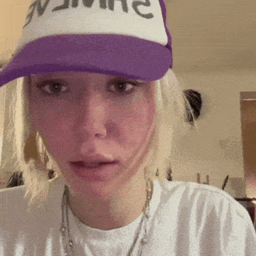 a woman wearing a white shirt and a purple hat with the word name on it looks at the camera