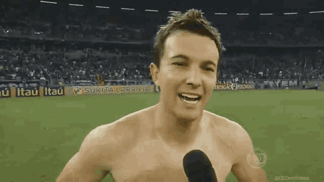 a shirtless man is talking into a microphone in front of a stadium with advertisements for sukita laranja