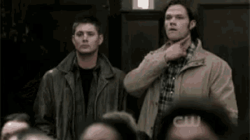 two men are standing next to each other in a crowd and one is pointing at his neck .