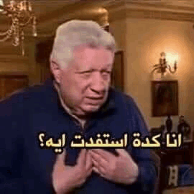 a man with gray hair is sitting in a living room with his hands on his chest and a caption in arabic .