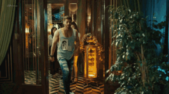 a man in a blue tank top is walking in a room with a woman behind him