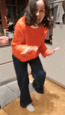 a girl in an orange sweater and blue jeans is dancing in a kitchen