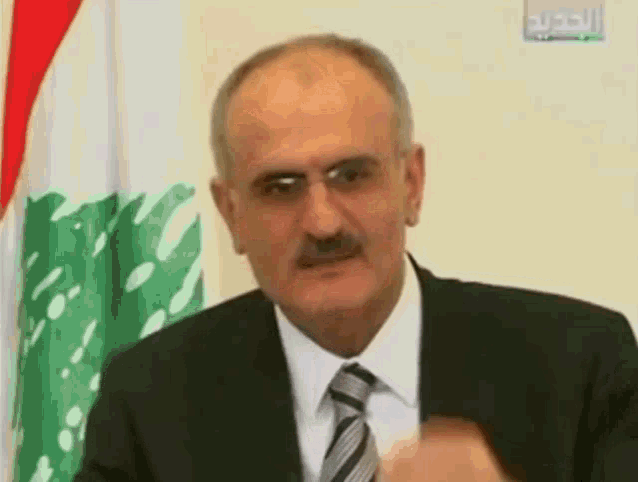 a man in a suit and tie is standing in front of a flag with arabic writing on it