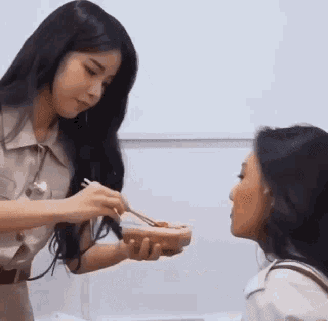 a woman is feeding another woman with chopsticks while holding a bowl of food .