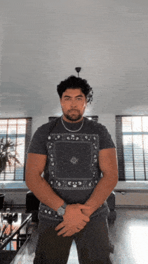 a man wearing a bandana t-shirt stands in a living room with his hands folded