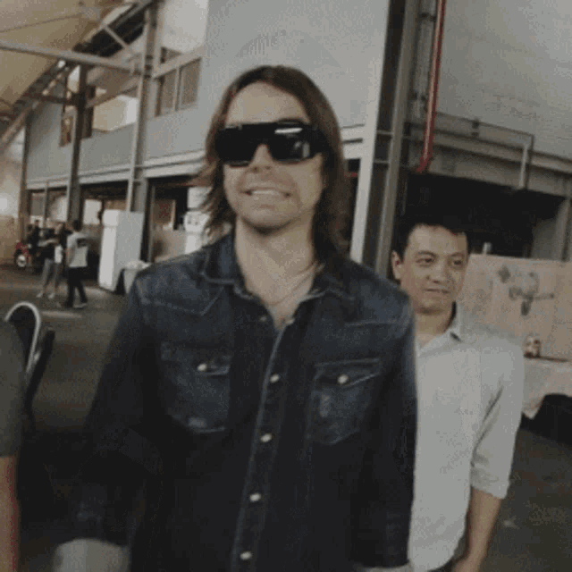 a man wearing sunglasses and a denim shirt stands in a crowd
