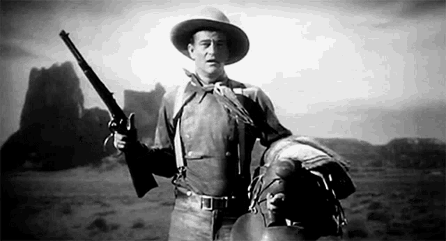 a man in a cowboy hat is holding a gun and a saddle in a black and white photo .