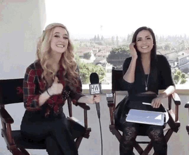 two women are sitting in director 's chairs and one is holding a microphone that says hw on it .
