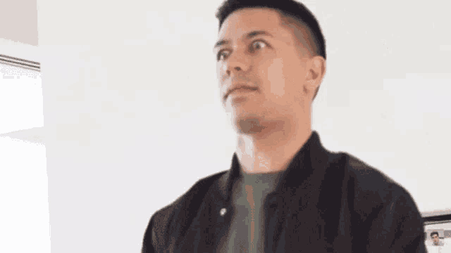 a man in a black jacket and green shirt is standing in front of a white wall .