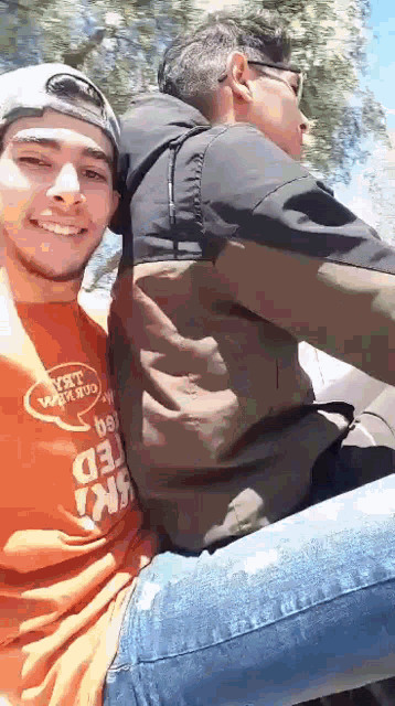 two men are sitting next to each other on a motorcycle . one of the men is wearing an orange shirt .