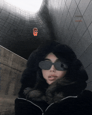a woman wearing sunglasses and a fur hood stands in front of a building with a g on it