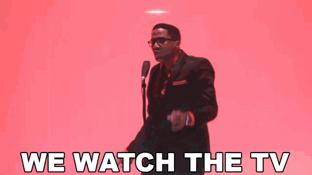 a man singing into a microphone with the words " we watch the tv " behind him