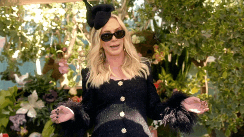 a woman wearing sunglasses and a black hat is standing in front of a garden