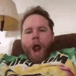 a man with a beard is yawning while laying on a couch wearing a green and yellow shirt .