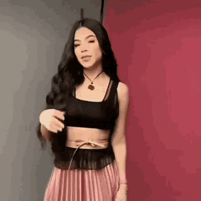a woman in a black crop top and a pink pleated skirt is standing in front of a red wall .