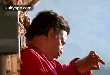 a man in a red shirt is smoking a cigarette while standing next to a building .