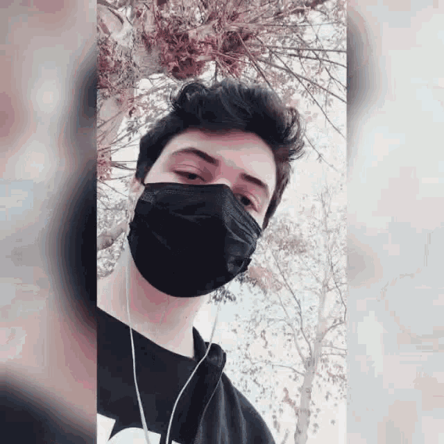 a young man wearing a black face mask and headphones stands in front of a tree .
