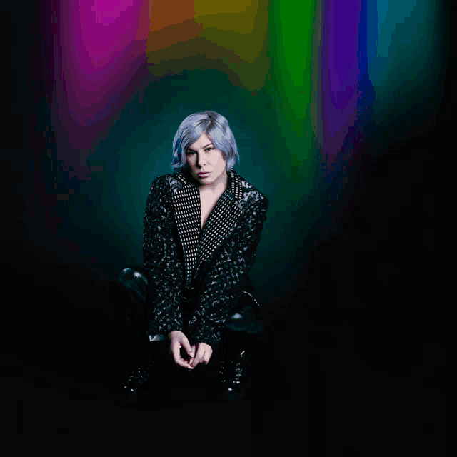 a woman with purple hair is sitting in front of a rainbow colored background