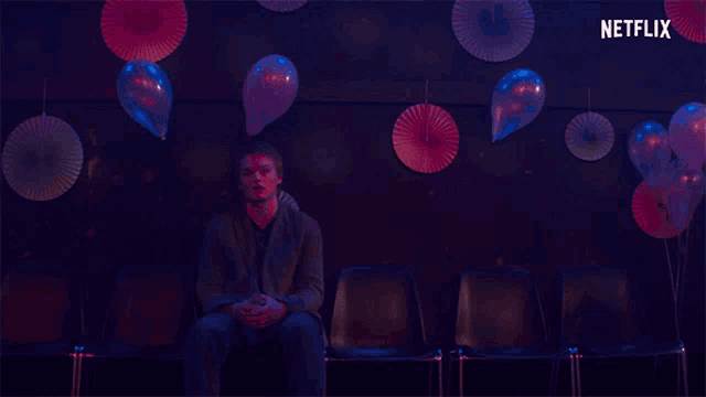 a man sits in a row of chairs with balloons and paper fans in the background and a netflix logo in the corner