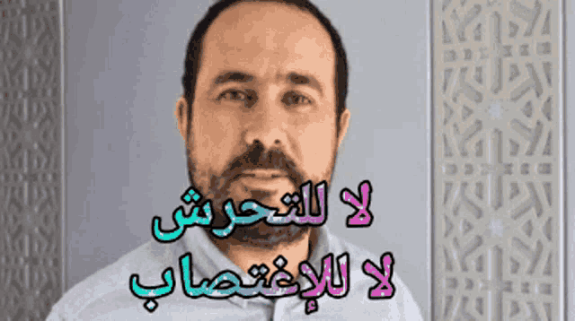 a man with a beard is standing in front of a wall that has arabic writing on it