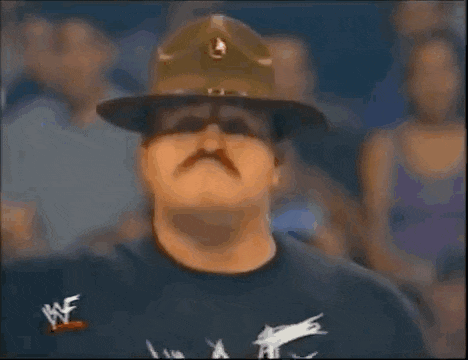 a man with a mustache wearing a military hat and a t-shirt is raising his fist in the air .