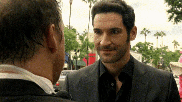 a man in a suit and black shirt is talking to another man in a suit and black shirt .