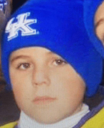 a young boy wearing a blue hat with the letter k on it looks at the camera