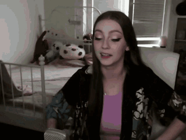 a woman wearing a purple top and a black jacket stands in front of a bed